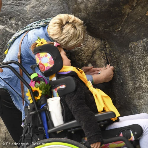 Enfant handicapé touchant la grotte de Lourdes avec accompagnateur.