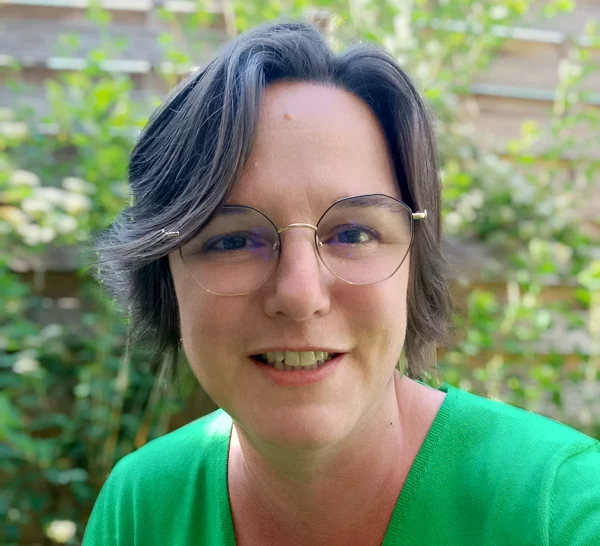 Portrait femme souriante avec lunettes, chemisier vert, extérieur.